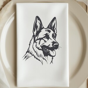May include: White cloth napkin featuring a black and white illustration of a German Shepherd dog's head. The dog is depicted in profile, with its tongue slightly out. The napkin is folded and placed on a beige plate.