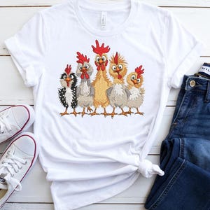 A white t-shirt with a graphic of five cartoon chickens, a pair of blue jeans, and white sneakers are arranged on a white wooden surface.