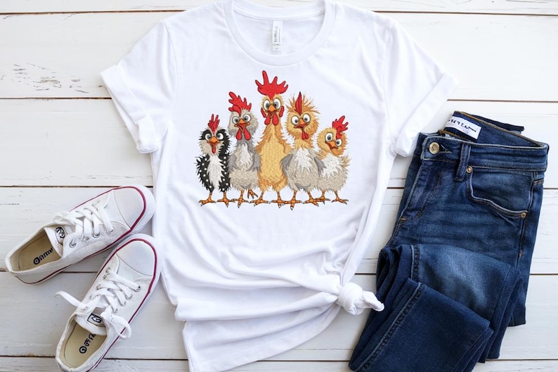 A white t-shirt with a graphic of five cartoon chickens, a pair of blue jeans, and white sneakers are arranged on a white wooden surface.