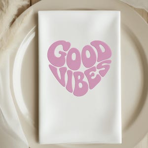 May include: White cloth napkin with a pink heart-shaped design that reads "GOOD VIBES". The napkin is folded and placed on a beige plate, suggesting a table setting. The design is in a retro font.