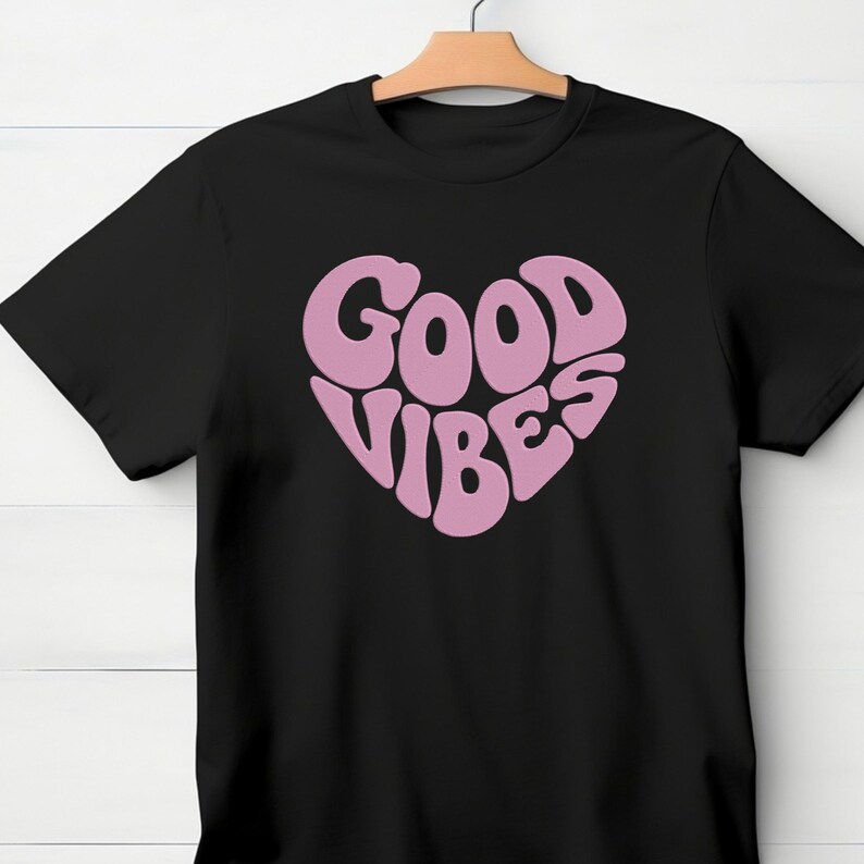 May include: Black t-shirt with a pink heart-shaped graphic that reads "GOOD VIBES" in a retro font. The shirt is hanging on a wooden hanger against a white background.
