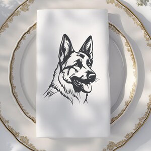 May include: A white cloth napkin featuring a black and white illustration of a German Shepherd dog's head. The napkin is folded and placed on a white plate with a gold rim. The dog's portrait shows its tongue and detailed fur.