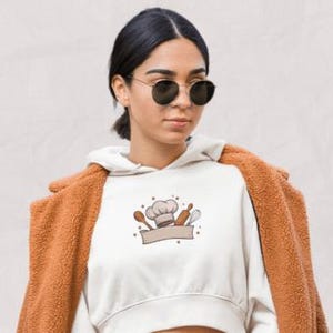 May include: A white cropped hoodie featuring a chef-themed graphic with a chef's hat, spoon, rolling pin, and banner. The person is wearing a brown coat and sunglasses. The background is a light beige color.