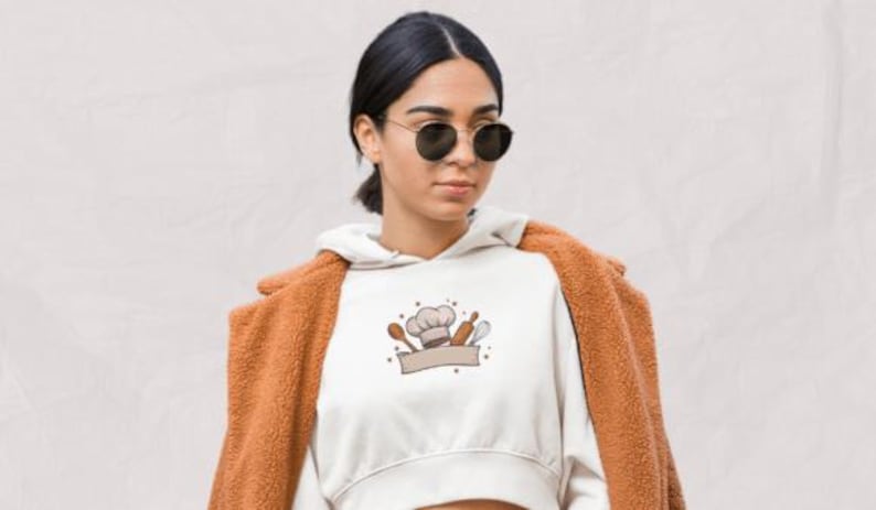 May include: A white cropped hoodie featuring a chef-themed graphic with a chef's hat, spoon, rolling pin, and banner. The person is wearing a brown coat and sunglasses. The background is a light beige color.