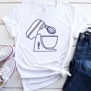 Puede incluir: Camiseta blanca con un dibujo lineal azul marino de una batidora de pie. La camiseta se combina con vaqueros azules y zapatillas blancas con ribetes rojos. Los artículos están colocados sobre una superficie de madera blanca.