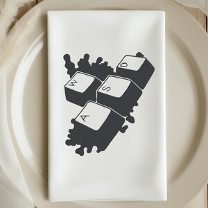 Peut inclure: Serviette en tissu blanche avec un motif noir de touches de clavier : W, A, S et D. Les touches sont regroup&eacute;es avec un fond sombre et abstrait. La serviette est pli&eacute;e et pos&eacute;e sur une assiette beige.