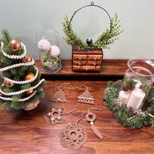 Puede incluir: Una exhibición festiva navideña con un pequeño árbol de Navidad decorado, adornos de macramé, velas y una corona decorativa. Los adornos son de varias formas y tamaños, con una paleta de colores neutros. La escena está sobre una superficie de madera.