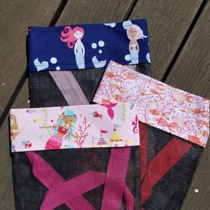 May include: Three mesh bags with different fabric designs. The top bag has a navy blue background with white and pink mermaids. The middle bag has a pink background with white and blue mermaids. The bottom bag has a red background with a white floral pattern.