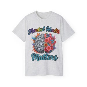 Könnte beinhalten: Hellgraues T-Shirt mit den Worten "Mental Health Matters" in blauer und violetter Schrift. Das Design zeigt ein in zwei Hälften geteiltes Gehirn, eine Seite ein detailliertes Gehirn, die andere ein farbenfrohes Blumenarrangement.