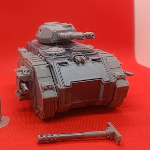 Pode incluir: Modelo de plástico cinzento de um tanque militar com uma grande torre e múltiplos componentes menores. O tanque está sobre uma superfície vermelha, com peças adicionais dispostas ao redor. O modelo é detalhado e parece ser uma réplica em miniatura.