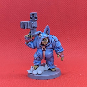 以下が含まれることがあります： 青いジャンプスーツとフードを着て、サイコロ型の武器を持っている生き物のミニチュアフィギュア。フィギュアは、小さな白い雲のような形のある灰色のベースに立っています。