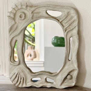 May include: An off-white ceramic mirror with a textured, organic frame. The frame has a sun-like design and irregular openings. The rectangular mirror with rounded corners sits on a wooden surface. The mirror's frame has a unique, sculptural quality.
