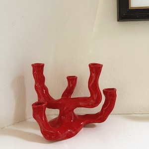 May include: A vibrant red ceramic candle holder with five openings. The sculptural piece has a unique, organic shape with curved, branch-like arms. The glossy finish and bold color make it a striking decorative accent.