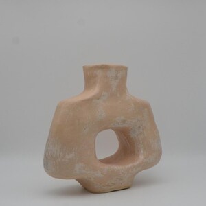May include: A peach-coloured, abstract ceramic vase with a large, circular opening. The vase has a textured surface with white accents, and a narrow neck. The design is modern and sculptural.
