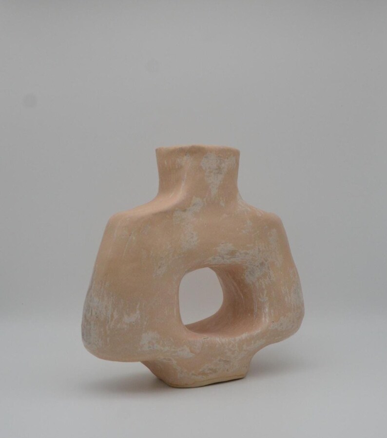 May include: A peach-coloured, abstract ceramic vase with a large, circular opening. The vase has a textured surface with white accents, and a narrow neck. The design is modern and sculptural.