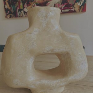 May include: A beige, abstract ceramic vase with a distinctive, open-centre design. The vase has a textured surface and a wide, rounded base. The top of the vase is shaped like a neck and shoulders. The vase is displayed on a wooden surface.