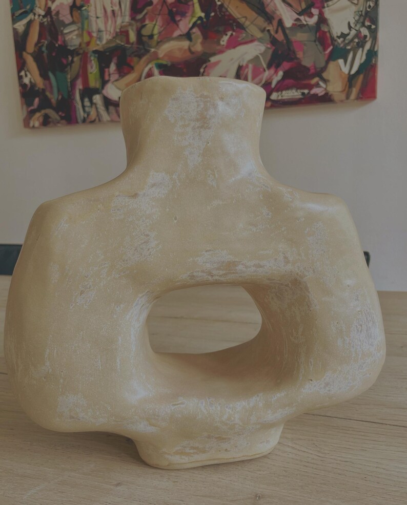 May include: A beige, abstract ceramic vase with a distinctive, open-centre design. The vase has a textured surface and a wide, rounded base. The top of the vase is shaped like a neck and shoulders. The vase is displayed on a wooden surface.