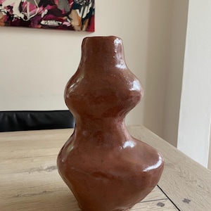 Könnte beinhalten: Eine braune Keramikvase mit einer einzigartigen, bauchigen Form. Die Vase hat einen schmalen Hals und einen breiten Boden, mit zwei abgerundeten Abschnitten in der Mitte. Die Oberfläche hat ein glänzendes Finish. Die Vase steht auf einem Holztisch.