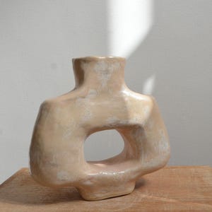 May include: A beige ceramic vase with a large, circular opening. The vase has a textured surface and a unique, organic shape. The vase is displayed on a wooden surface.