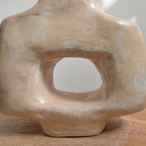 May include: A beige ceramic sculpture of a torso with a large, oval-shaped opening in the centre. The sculpture has a smooth, glossy finish with subtle variations in colour and texture, suggesting a handmade quality. The piece is displayed on a wooden surface.