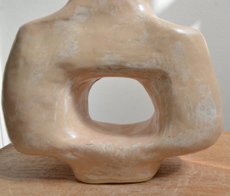 May include: A beige ceramic sculpture of a torso with a large, oval-shaped opening in the centre. The sculpture has a smooth, glossy finish with subtle variations in colour and texture, suggesting a handmade quality. The piece is displayed on a wooden surface.
