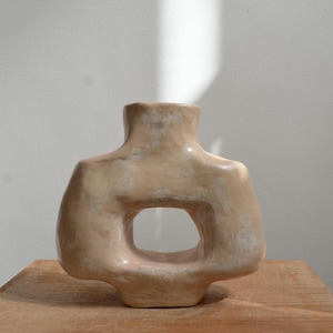 May include: A beige ceramic vase with a distinctive open-centre design. The vase has a rounded shape with a narrow neck and a wide base. The surface has a slightly textured appearance.