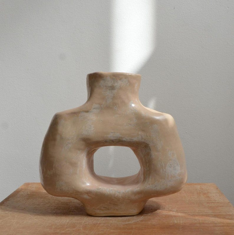 May include: A beige ceramic vase with a distinctive open-centre design. The vase has a rounded shape with a narrow neck and a wide base. The surface has a slightly textured appearance.