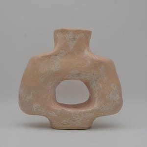 May include: A peach-coloured ceramic vase with a large, oval-shaped opening. The vase has a unique, organic form with a textured surface and a neutral background. The design is modern and minimalist.
