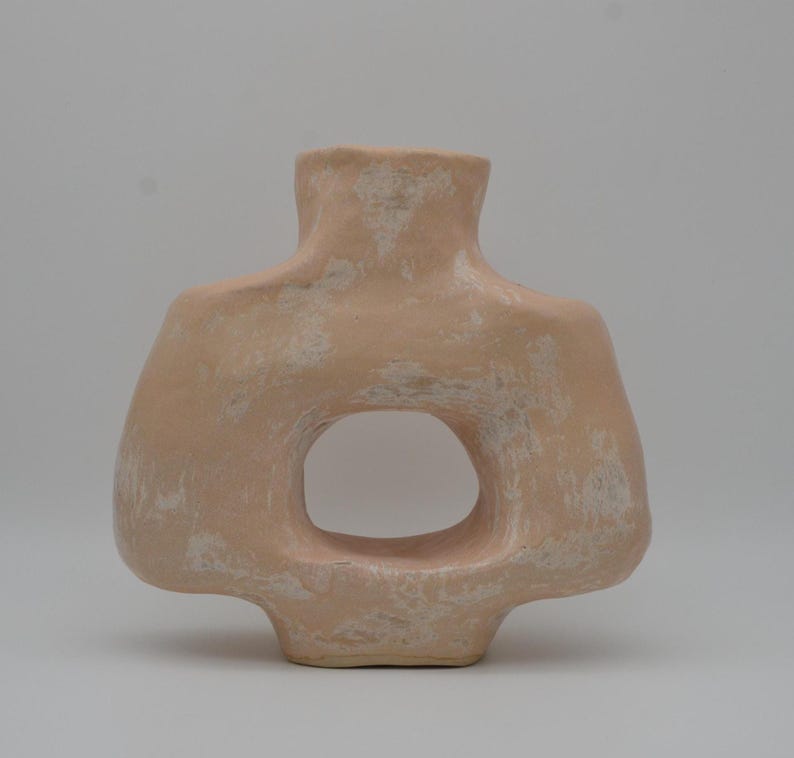 May include: A peach-coloured ceramic vase with a large, oval-shaped opening. The vase has a unique, organic form with a textured surface and a neutral background. The design is modern and minimalist.