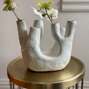 Könnte beinhalten: Weiße Keramikvase mit einer einzigartigen, organischen Form und mehreren Öffnungen für Blumen. Die Vase steht auf einem goldenen Metallständer.