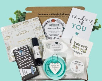 May include: A gift box with various self-care items. Includes a chocolate bar, a rollerball bottle labeled "peace", bath bombs, a card that says "thinking of you", and a heart-shaped sign that says "Sending you the biggest hug".