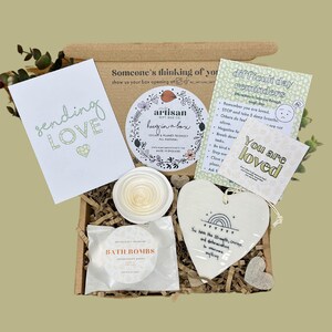 Könnte beinhalten: Eine braune Pappbox gefüllt mit Selbstpflegeartikeln. Die Box enthält eine Karte mit der Aufschrift "sending love", ein weißes Keramikherz mit dem Text "You are loved", eine weiße Keramikschale mit einem Spiralmuster, eine weiße Badebombe und eine weiße Karte mit dem Text "difficult day reminders".