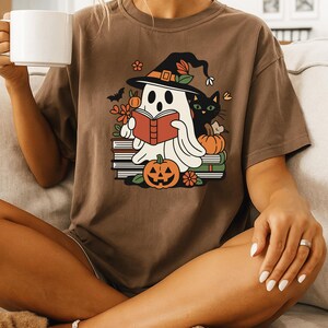 Può includere: T-shirt marrone con grafica a tema Halloween. Il design presenta un fantasma che legge un libro, un gatto nero, zucche e fiori. Il fantasma indossa un cappello da strega. La maglietta è di vestibilità ampia.