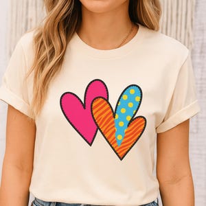 Può includere: T-shirt color crema con tre cuori colorati. I cuori presentano motivi unici: un cuore rosa pieno, un cuore a righe arancioni e gialle e un cuore blu a pois gialli. Ogni cuore è delineato da una cucitura nera.