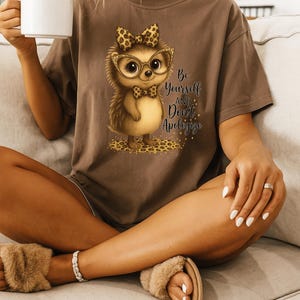 Può includere: T-shirt marrone con un riccio dei cartoni animati con occhiali e papillon, con la scritta "Be Yourself and Don't Apologize". Il riccio è su una stampa leopardata. La persona indossa un braccialetto e delle pantofole.