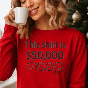 May include: A red long-sleeve shirt with the text "This shirt is $50,000" in black. Below the text is a black decorative design and the word "Shisgama" in script. The shirt is being worn by a person holding a white mug.