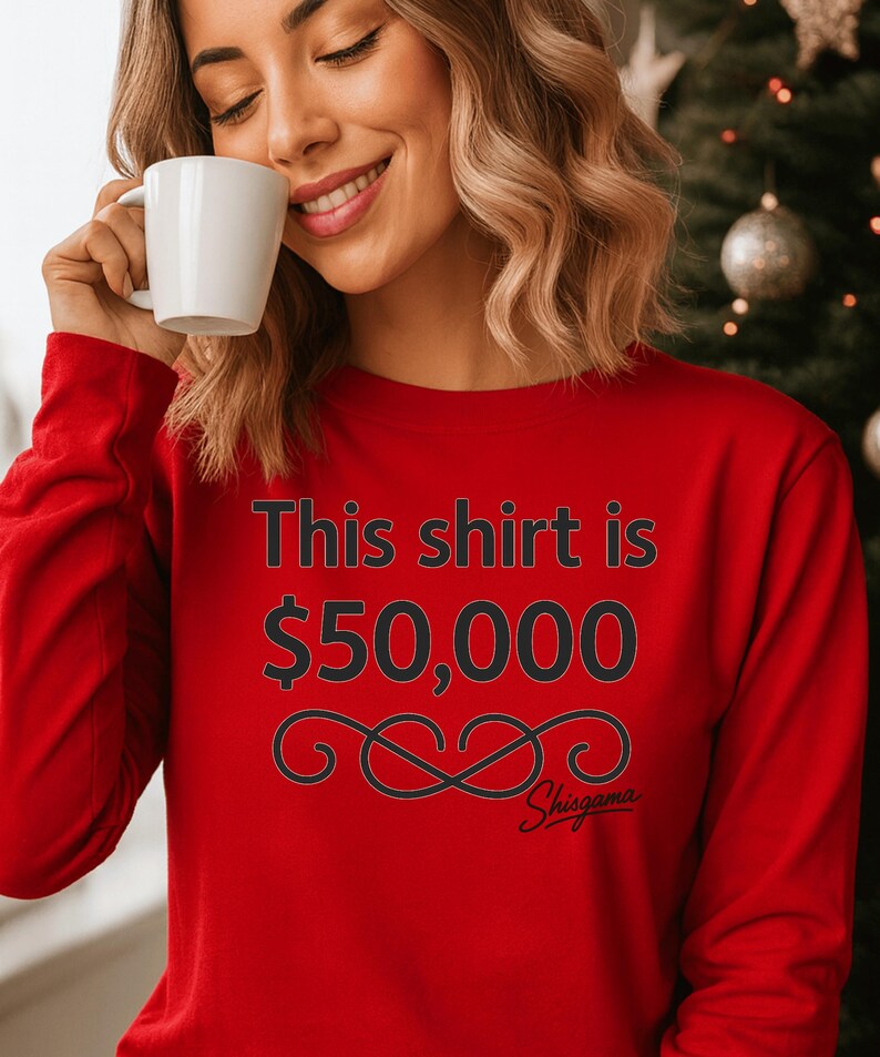May include: A red long-sleeve shirt with the text "This shirt is $50,000" in black. Below the text is a black decorative design and the word "Shisgama" in script. The shirt is being worn by a person holding a white mug.