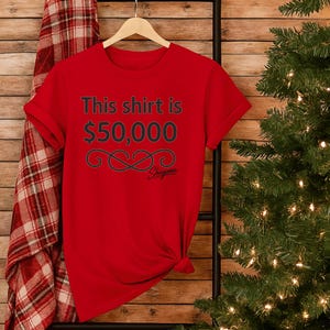 May include: A red t-shirt with the text "This shirt is $50,000" in black. Below the text is a decorative infinity symbol and the brand name "Shigama". The shirt is hanging on a wooden hanger.