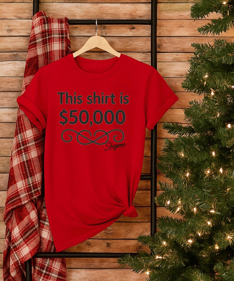 May include: A red t-shirt with the text "This shirt is $50,000" in black. Below the text is a decorative infinity symbol and the brand name "Shigama". The shirt is hanging on a wooden hanger.