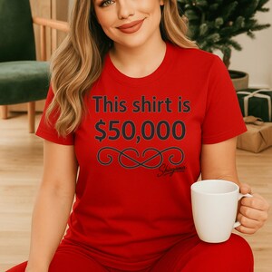May include: Red t-shirt with the text "This shirt is $50,000" in black, along with a decorative swirl and the brand name "Shugama". The shirt is part of a red outfit and is paired with a white mug.