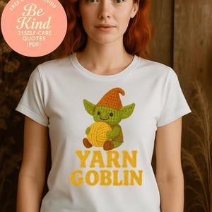 May include: White t-shirt featuring a cartoon goblin holding a ball of yarn. The goblin is green with an orange hat and brown shoes. The text "YARN GOBLIN" is in yellow. A pink circle with text is in the upper left corner.