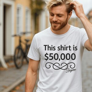 May include: White crew neck t-shirt with the text "This shirt is $50,000" in black. Below the text is a decorative swirl design and the brand name "Shisgama". The shirt is being worn.