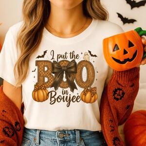 Può includere: T-shirt bianca con grafica a tema Halloween. Il design include la scritta "I put the BOO in Boujee" con un fiocco nero, zucche, pipistrelli e un fantasma. La persona tiene in mano una tazza a forma di zucca arancione.