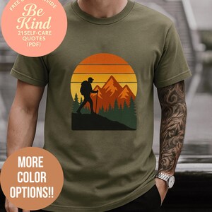 Può includere: T-shirt verde oliva con un design a silhouette di escursionista e tramonto montano nei colori arancione, giallo e verde. La maglietta presenta il testo "MORE COLOR OPTIONS!!" e "Be Kind".