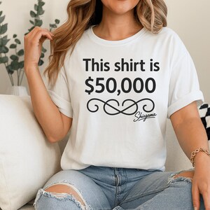 May include: White t-shirt with the text "This shirt is $50,000" in black, along with a decorative infinity symbol and the brand name "Shisgama". The shirt is worn with blue jeans.
