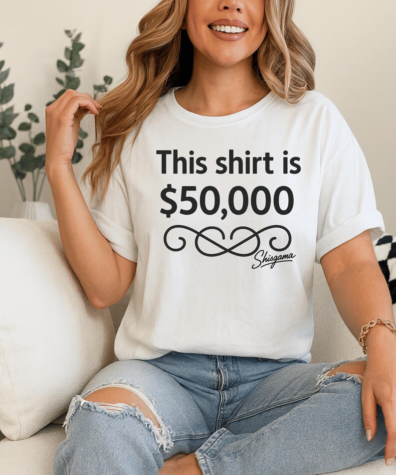 May include: White t-shirt with the text "This shirt is $50,000" in black, along with a decorative infinity symbol and the brand name "Shisgama". The shirt is worn with blue jeans.