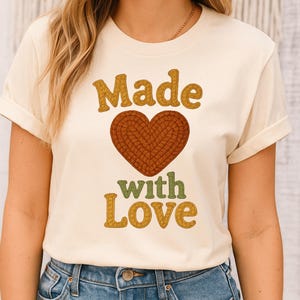 Può includere: T-shirt color crema con la scritta "Made with Love" in un font retrò. La parola "Made" è di colore giallo senape, e "with Love" è verde e giallo. Un cuore marrone è al centro.