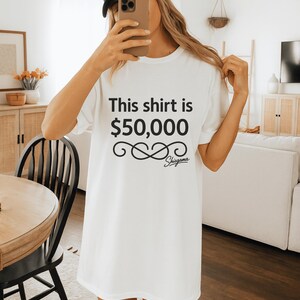 May include: White t-shirt with the text "This shirt is $50,000" in black, along with a decorative infinity symbol and the brand name "Shigama". The shirt is a simple, casual style.