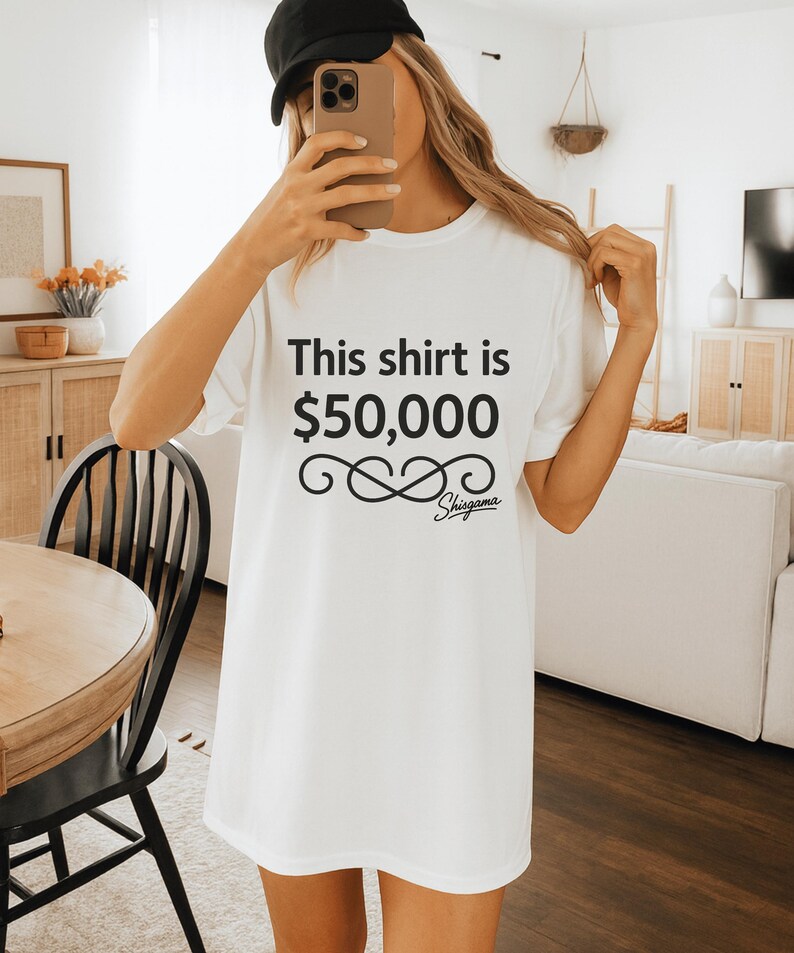 May include: White t-shirt with the text "This shirt is $50,000" in black, along with a decorative infinity symbol and the brand name "Shigama". The shirt is a simple, casual style.