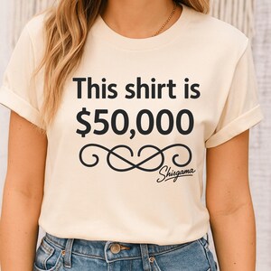 May include: Cream-colored t-shirt with the text "This shirt is $50,000" in black, along with a decorative swirl and the word "Shisgama" in script. The shirt has rolled-up sleeves and is worn with blue jeans.